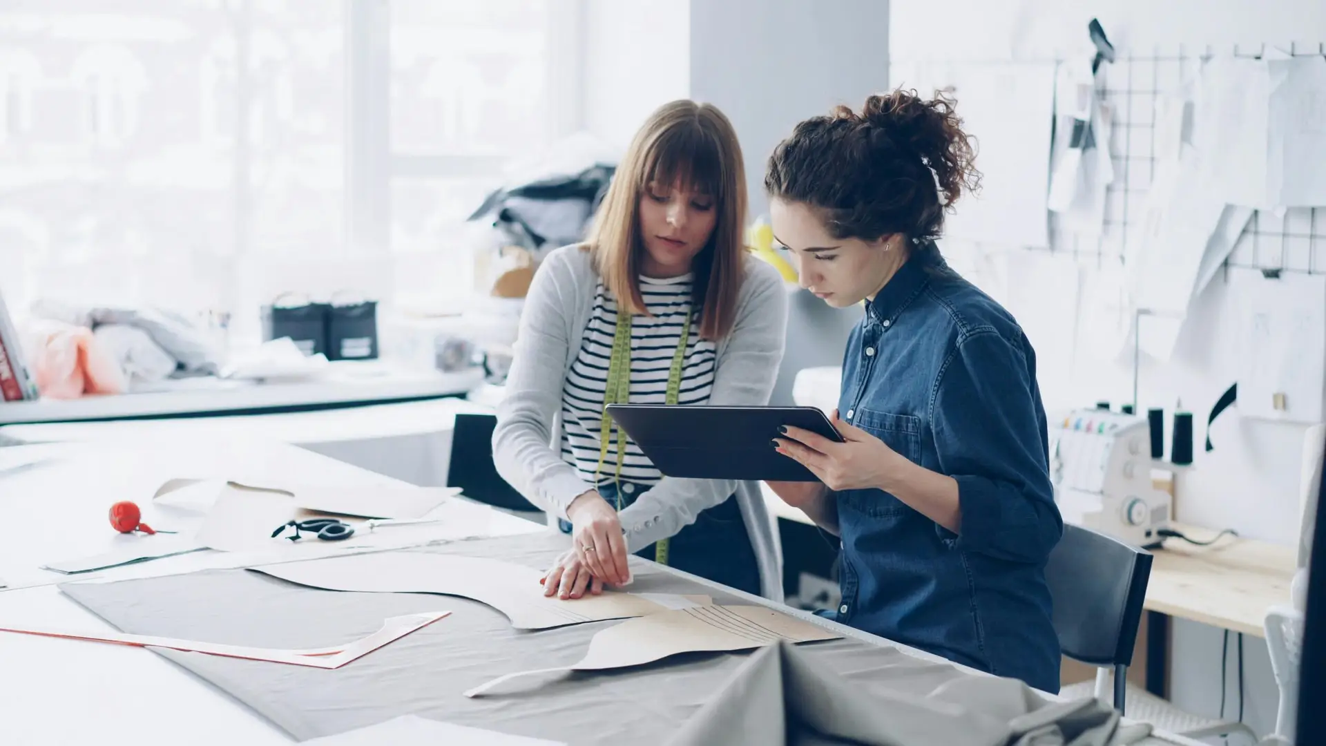 Two female fashion designers working together over fabric patterns in a bright studio.